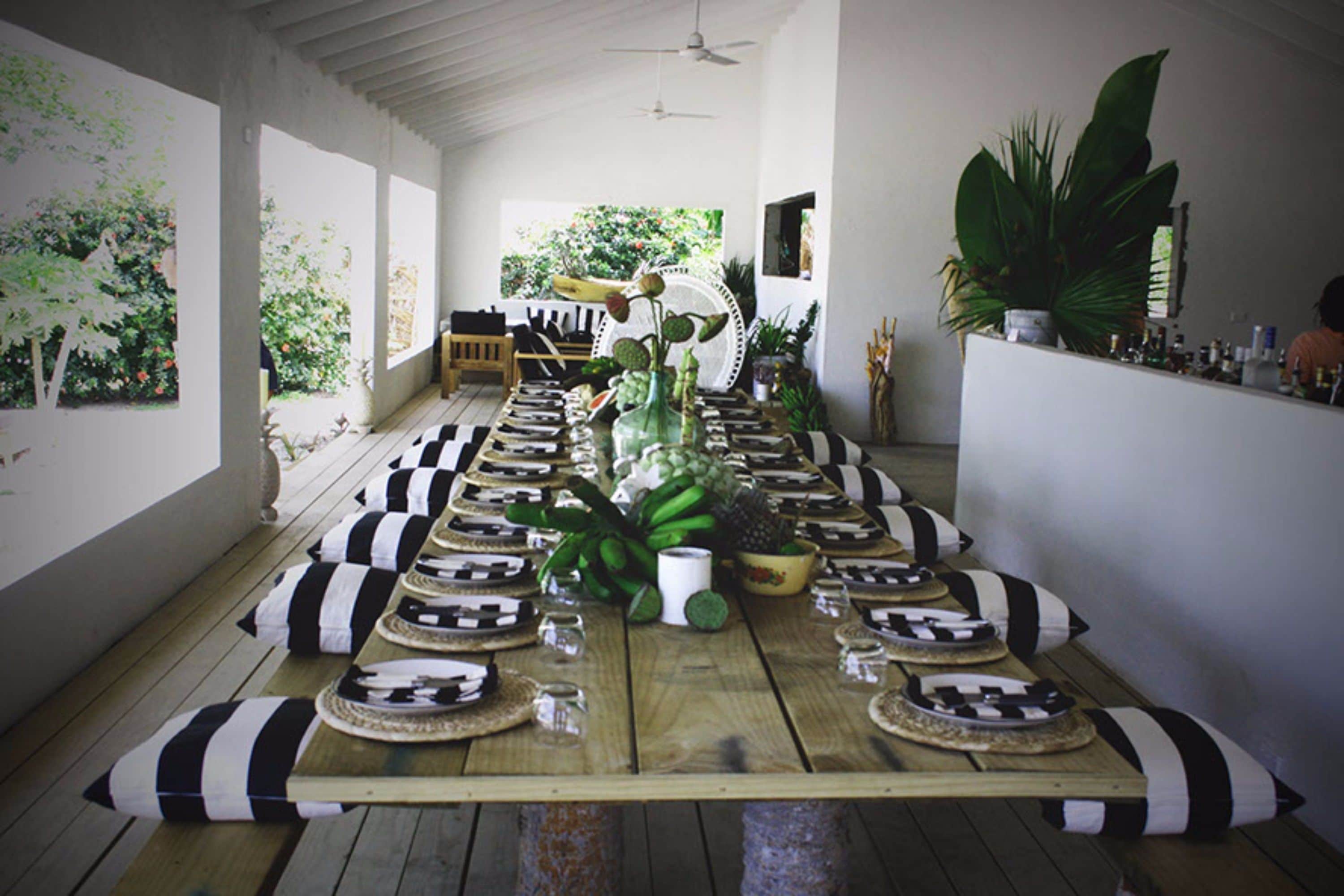 A long wooden table set for a meal with black and white striped cushions, surrounded by potted plants and a view of greenery outside.