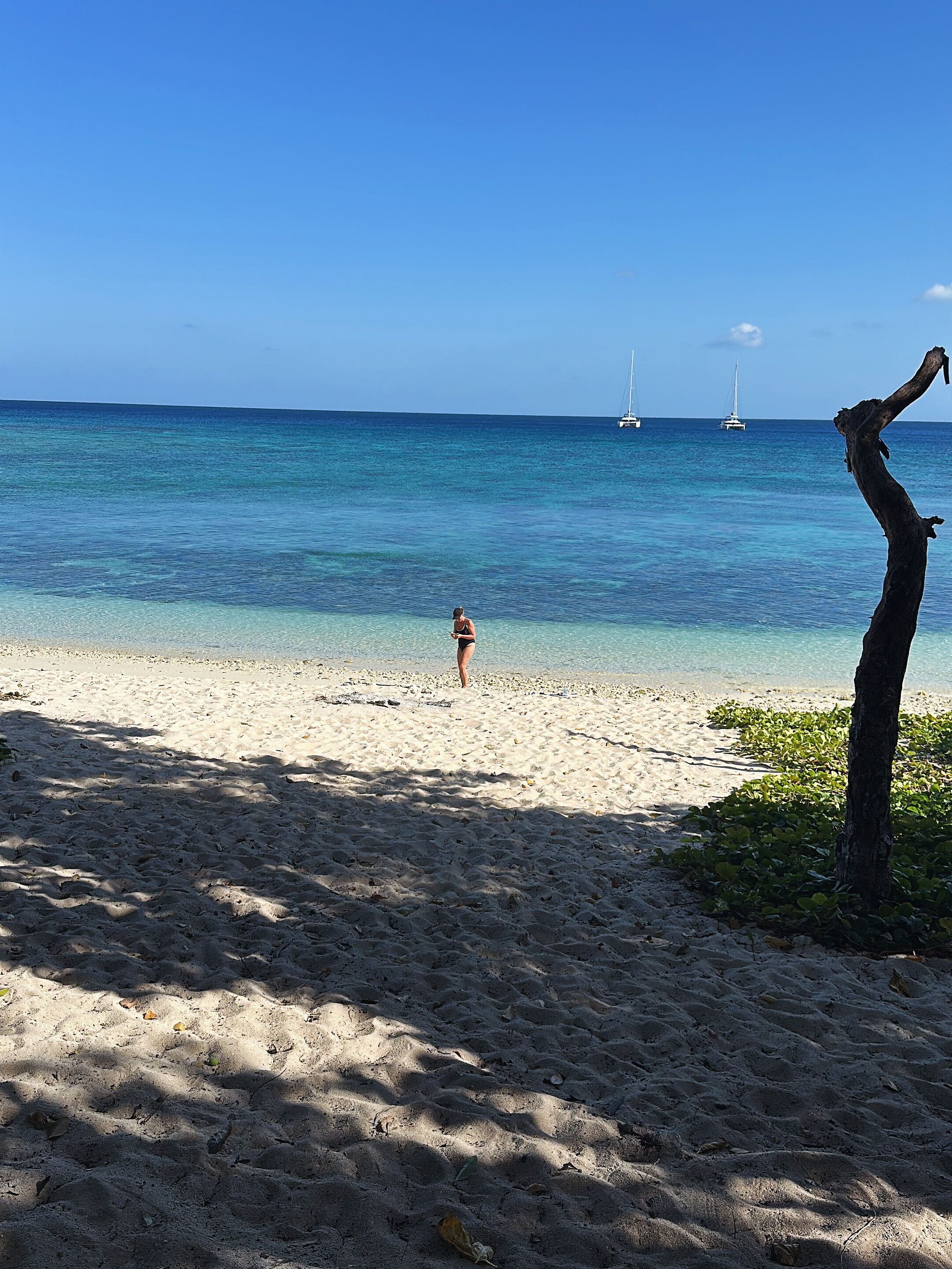 A beach with clear blue water, a person walking, and sailboats in the distance.