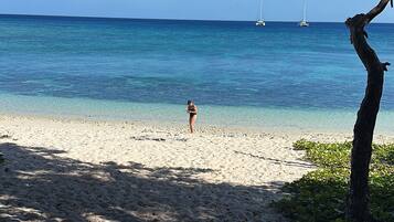 A beach with clear blue water, a person walking, and sailboats in the distance.