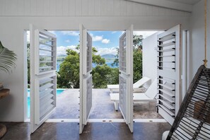 Eine offene Tür, die auf einen Balkon mit Pool, weißen Möbeln und einem Hängesessel führt.