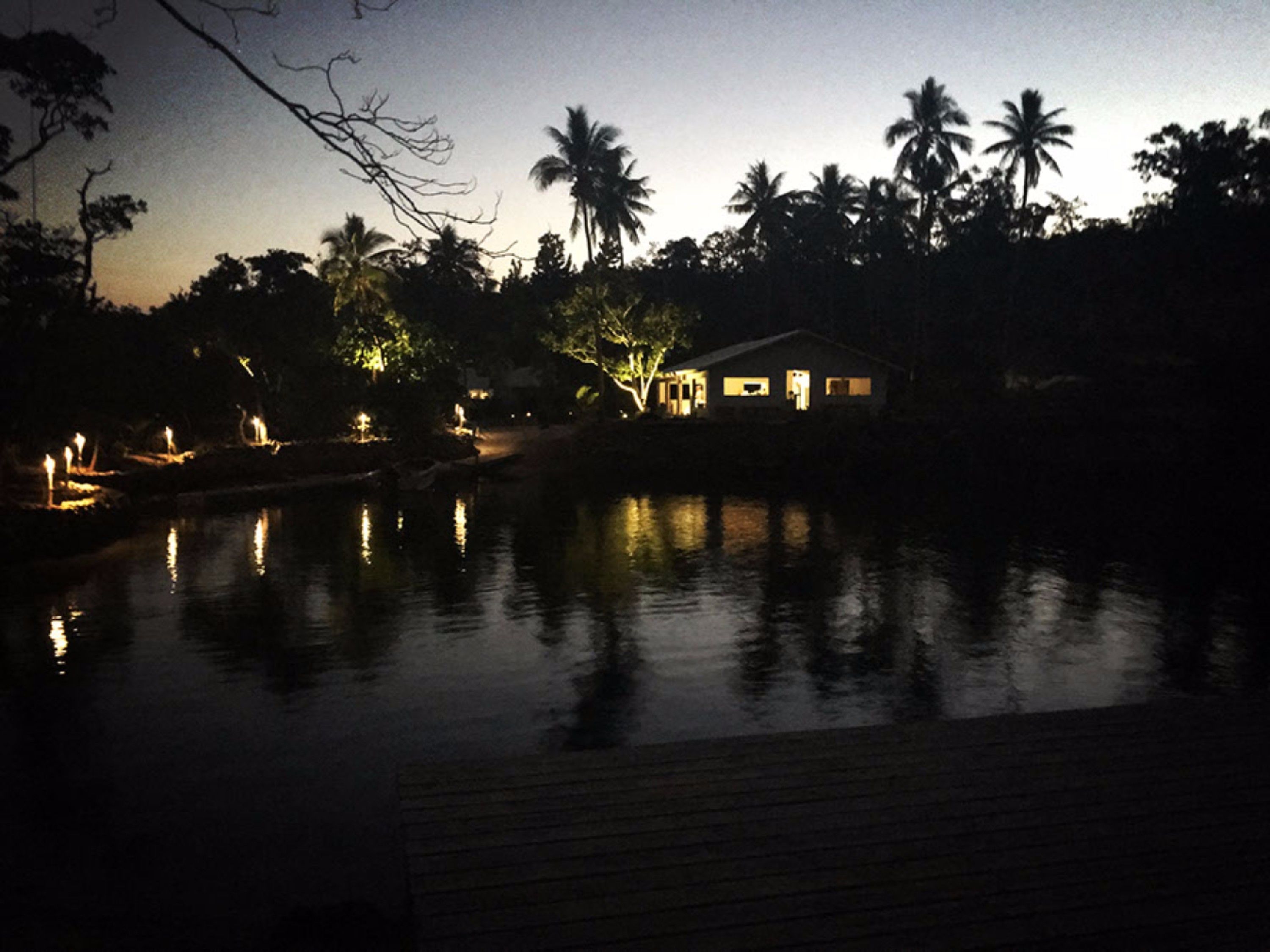 Escena nocturna con una masa de agua tranquila que refleja luces, rodeada de palmeras y una casa con ventanas iluminadas.