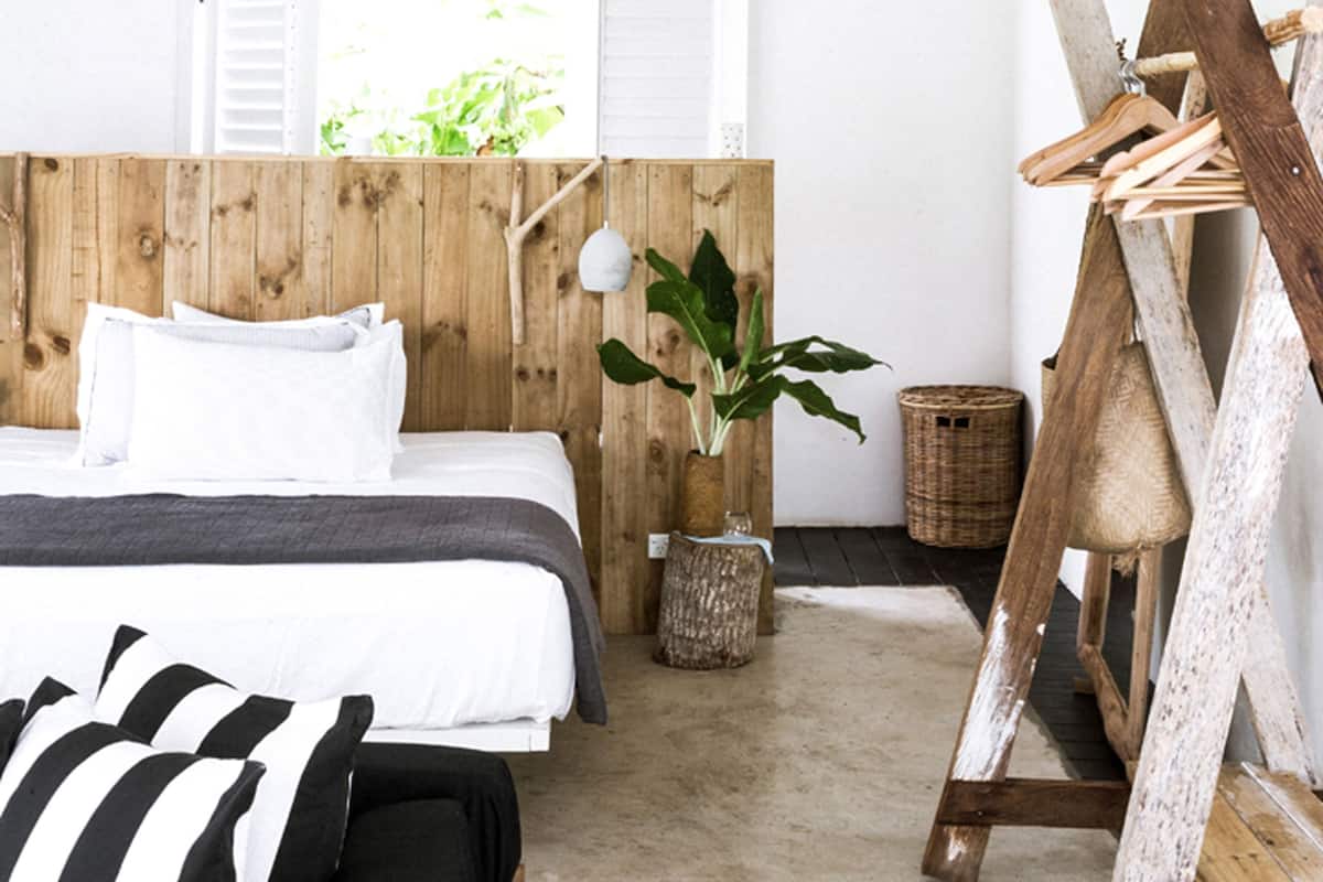 Un dormitorio con cabecera de madera, cama con ropa de cama blanca, almohada a rayas negras y blancas, una planta en maceta y un caballete de madera.
