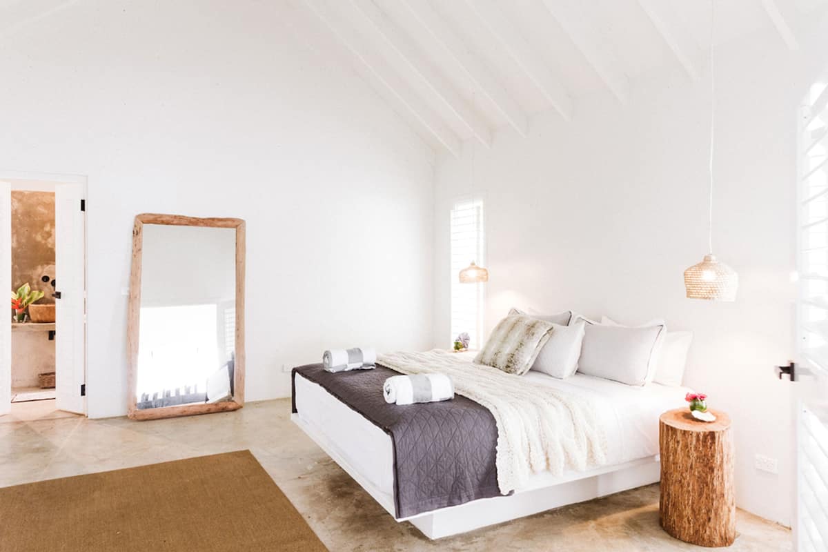 Un dormitorio con una cama grande, un espejo con marco de madera, una mesita de noche redonda de madera y una lámpara colgante.