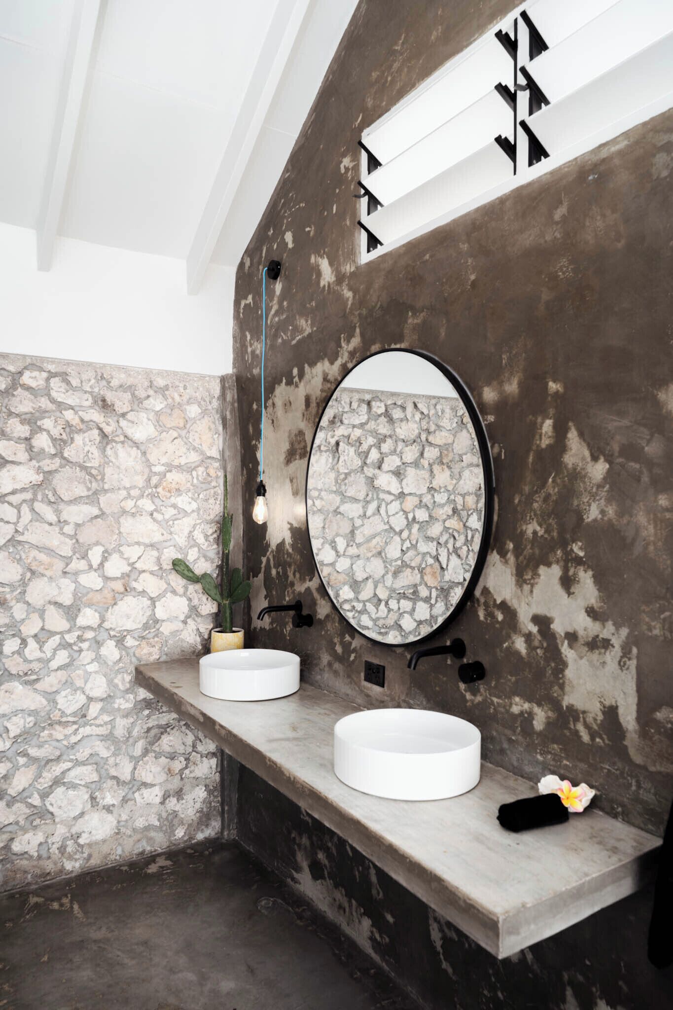 Un baño con pared de piedra, dos lavamanos, un espejo redondo y una planta en maceta.