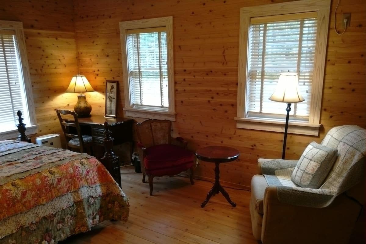 A cozy room with a bed, a desk, a chair, and a sofa. There are windows with blinds and a lamp on the desk.