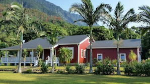 A red building with a white porch, surrounded by palm trees and a mountainous backdrop. - Cottage HANA MANA (Yakushima)