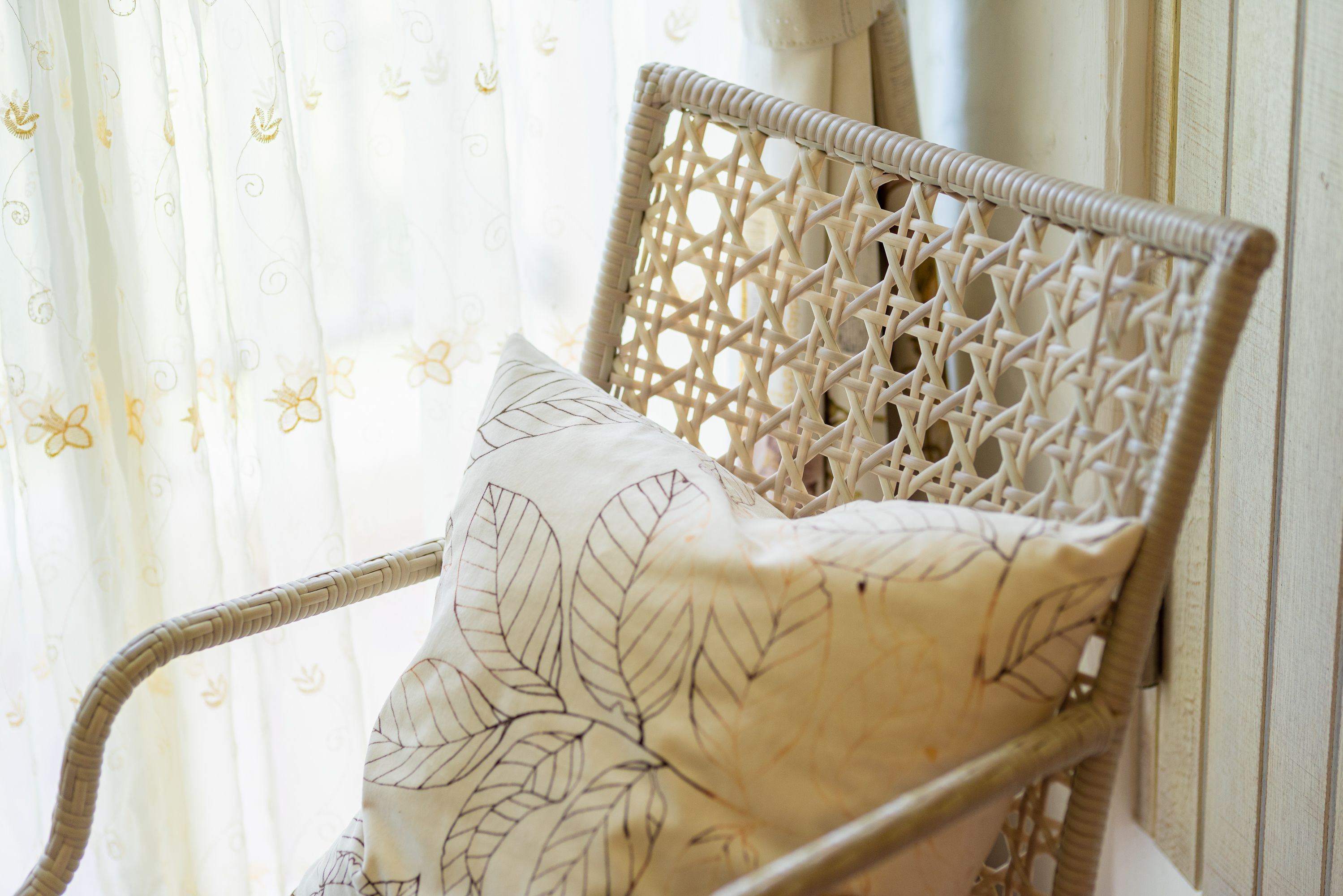 A wicker chair with a leaf-patterned cushion, positioned near a window with sheer curtains.