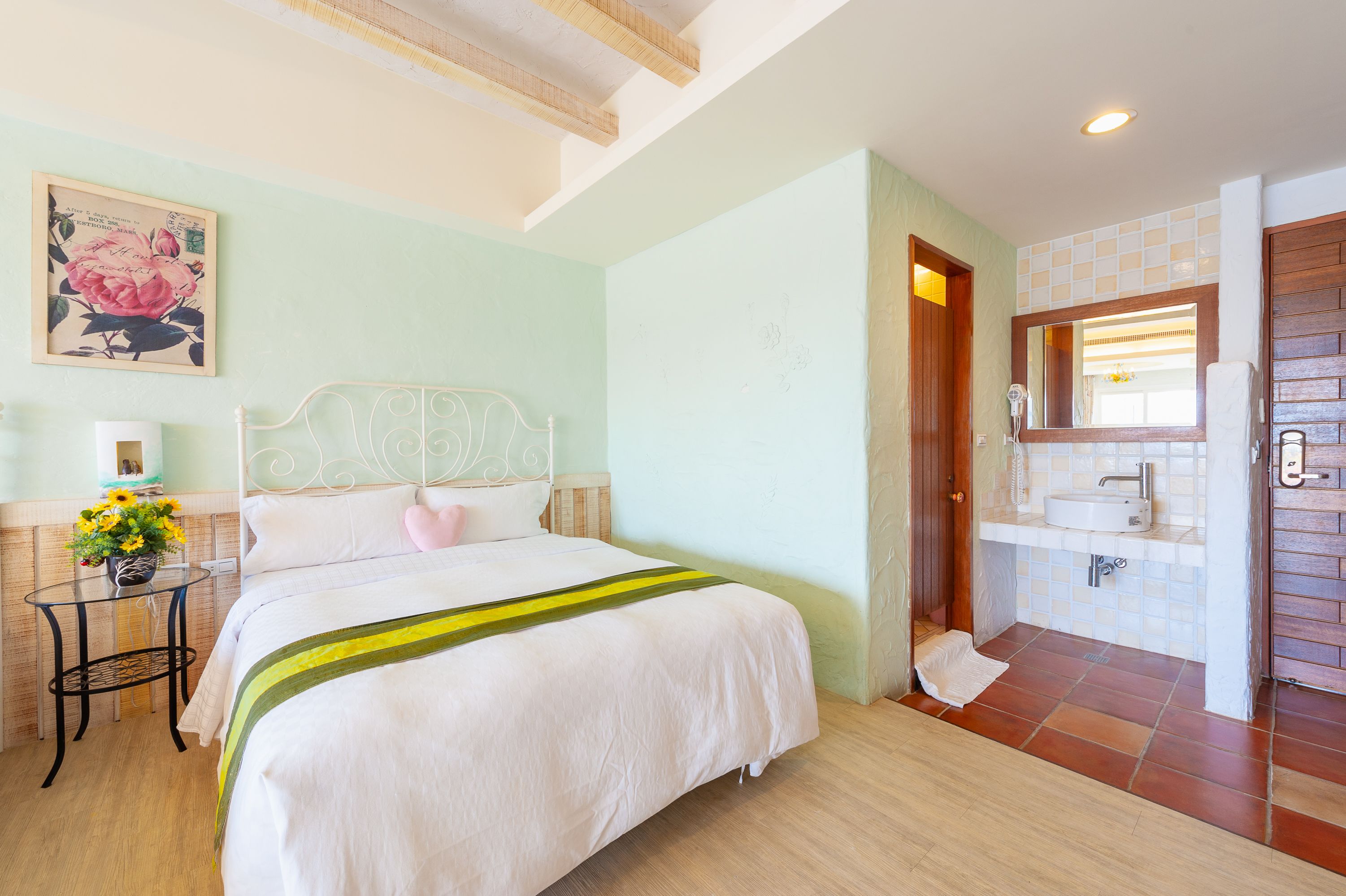 Un dormitorio con cama, mesita de noche con un florero y baño con lavamanos y espejo.