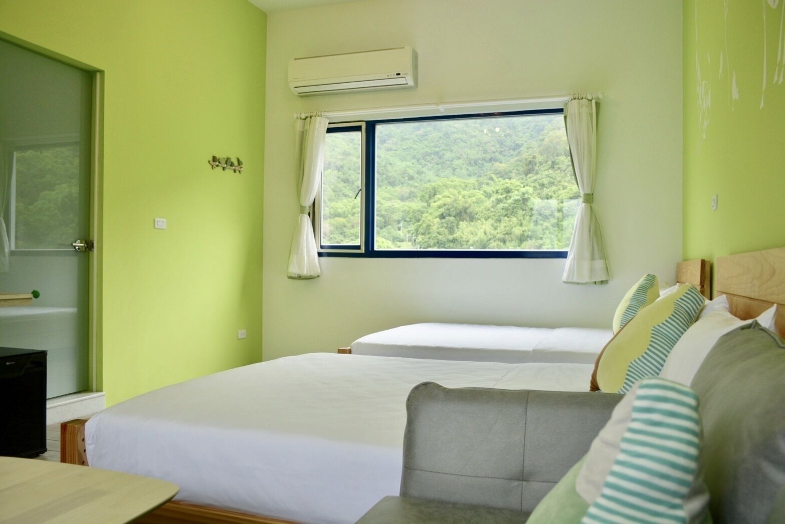 Une chambre avec deux lits, une fenêtre avec des rideaux, une petite table et un appareil de climatisation.