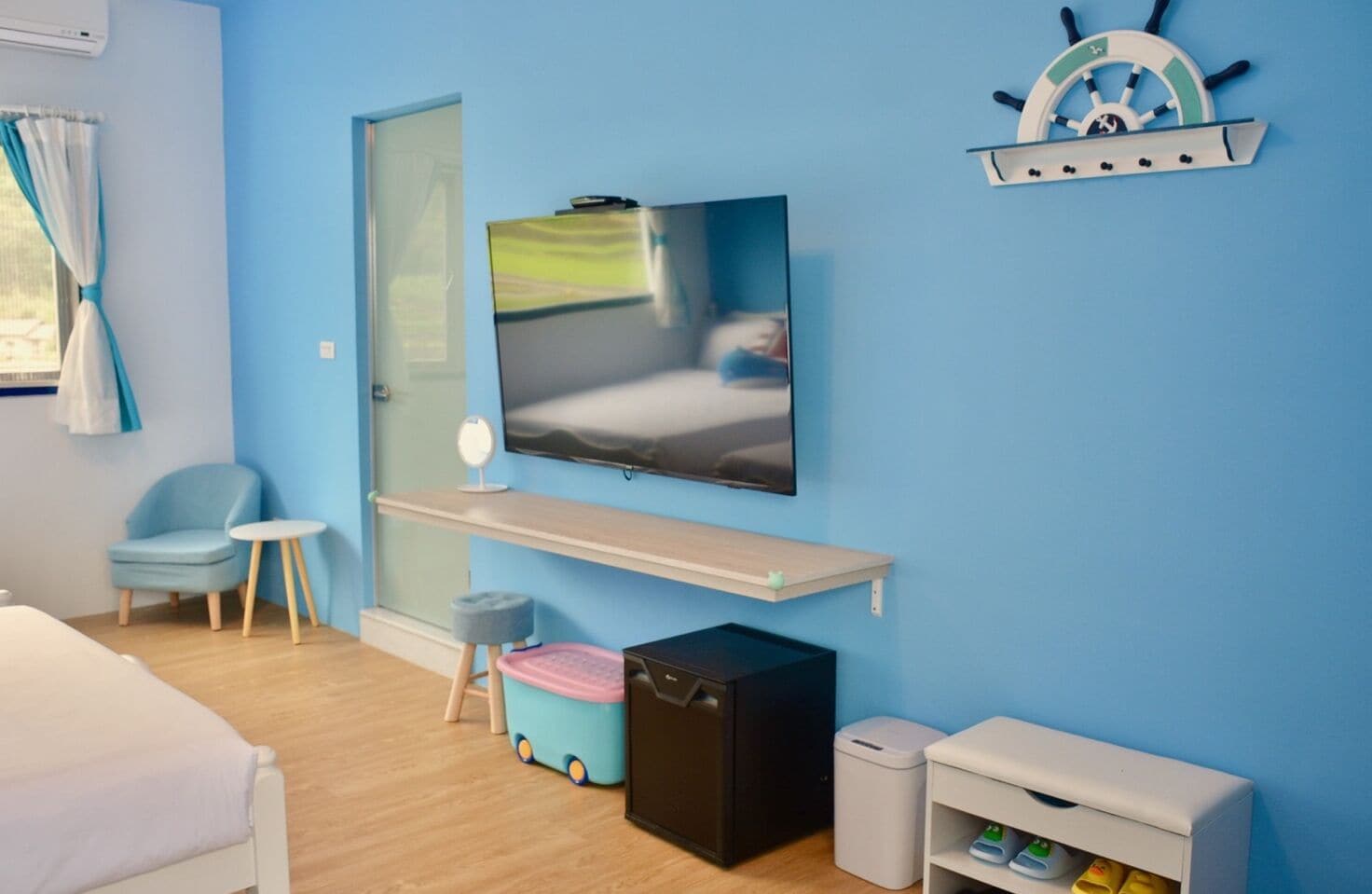 A room with a bed, a TV mounted on the wall, a small desk, a chair, a storage unit, and a shelf with a ship's wheel decoration.