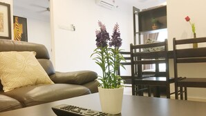 A living room with a brown leather sofa, a white cushion, a remote control, and a potted plant with purple flowers. - Mansion One Bedroom Suite (George Town)