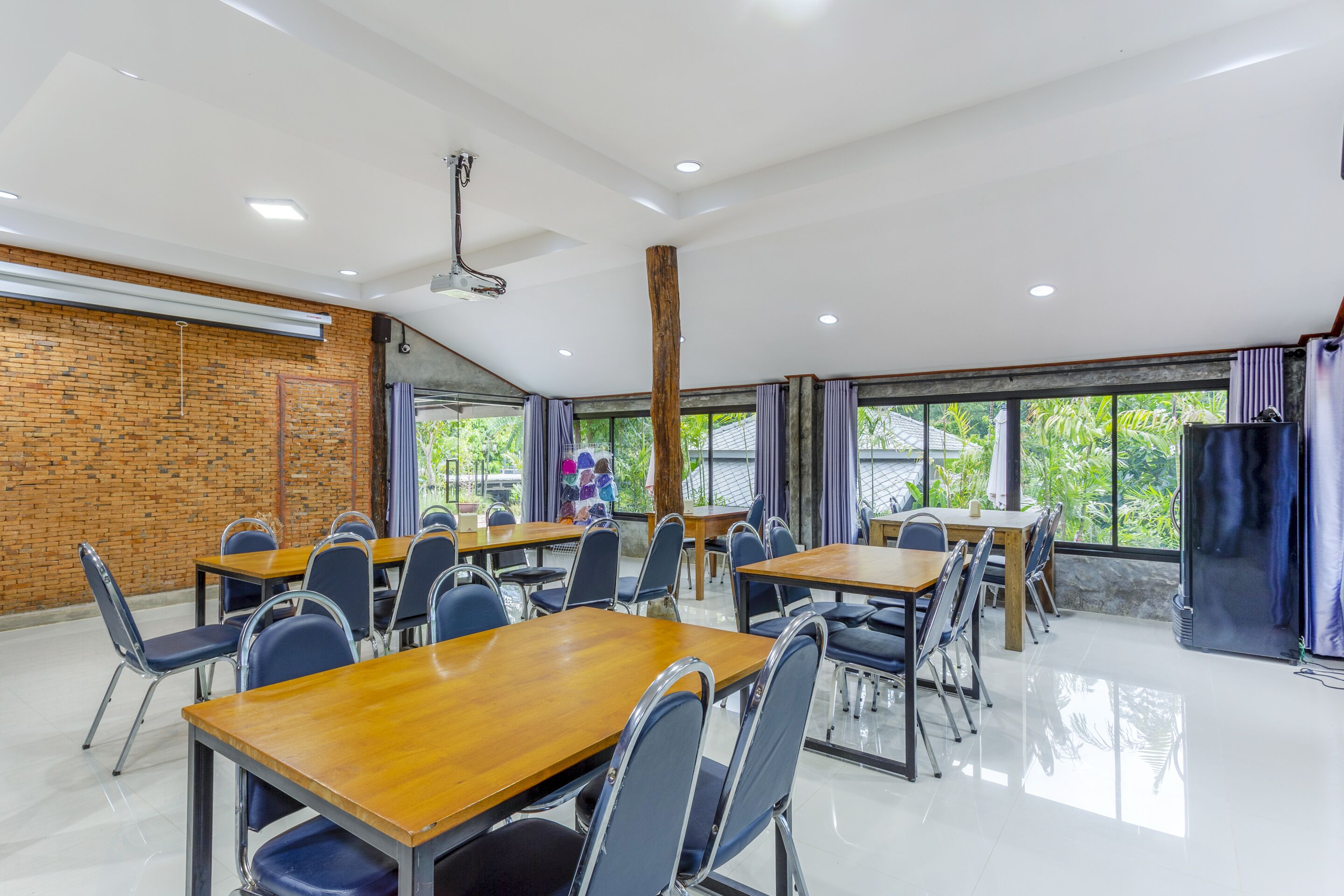 Une salle de conférence dotée de plusieurs tables et chaises, de grandes fenêtres avec des rideaux, d’un mur en briques et d’un ventilateur de plafond.