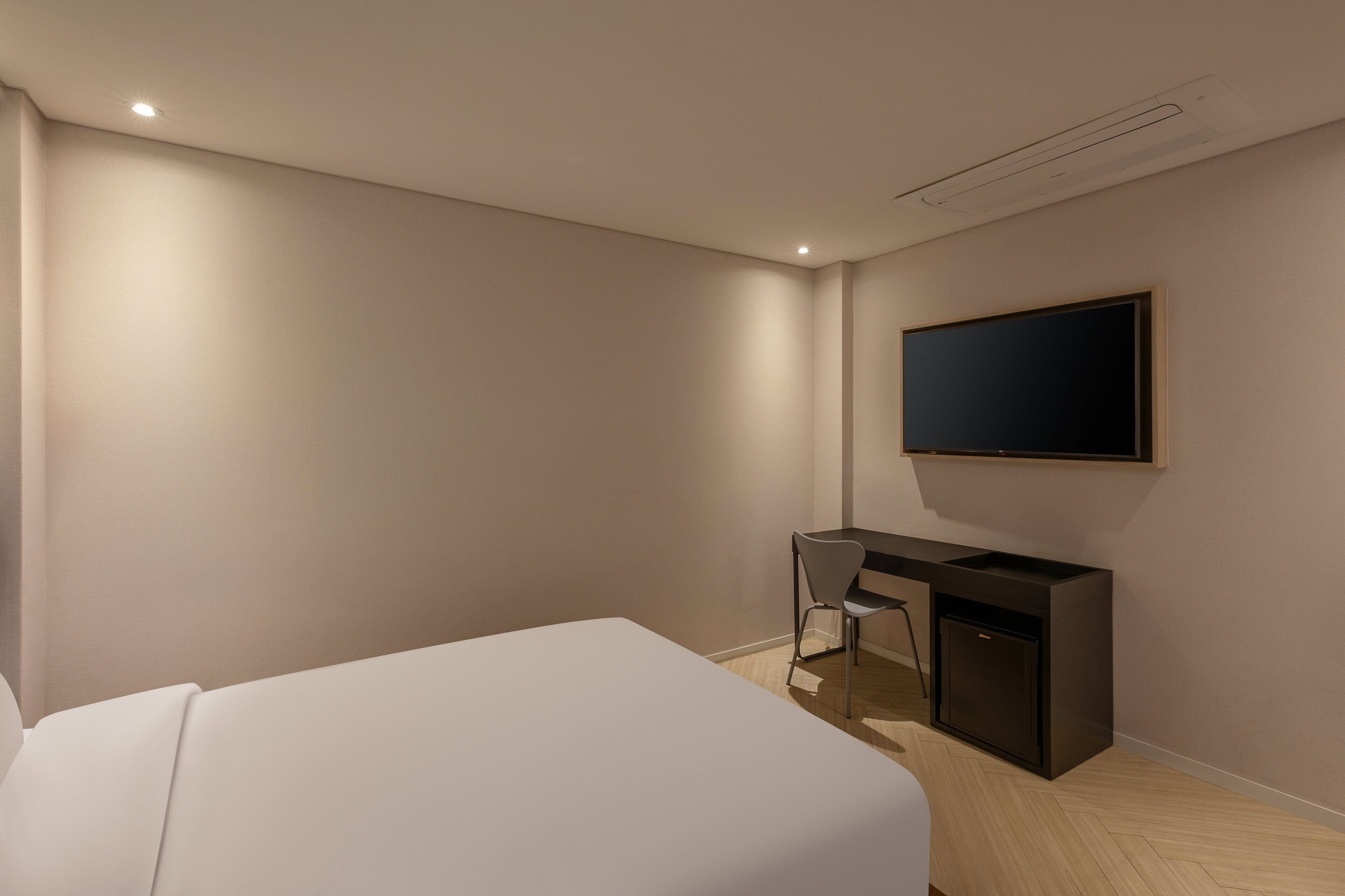 A hotel room with a bed, a desk, a chair, a television mounted on the wall, and a ceiling vent.