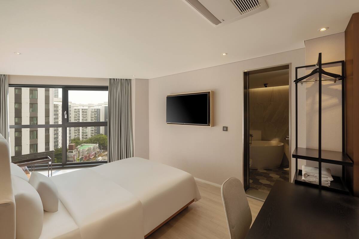 A modern hotel room with a large bed, a TV mounted on the wall, a shower area, and a view of the cityscape through the window.