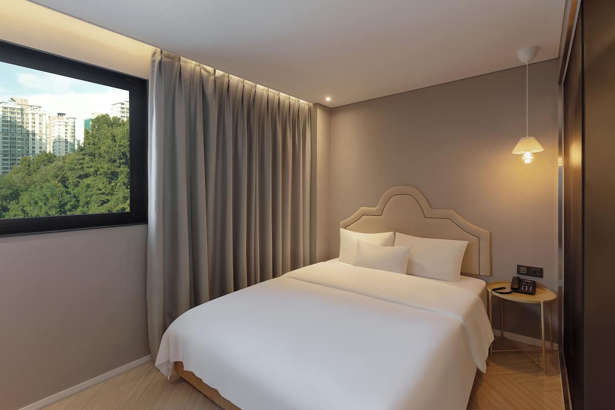 A hotel room with a large bed, a bedside table with a phone, a window with curtains, and a view of buildings and trees.