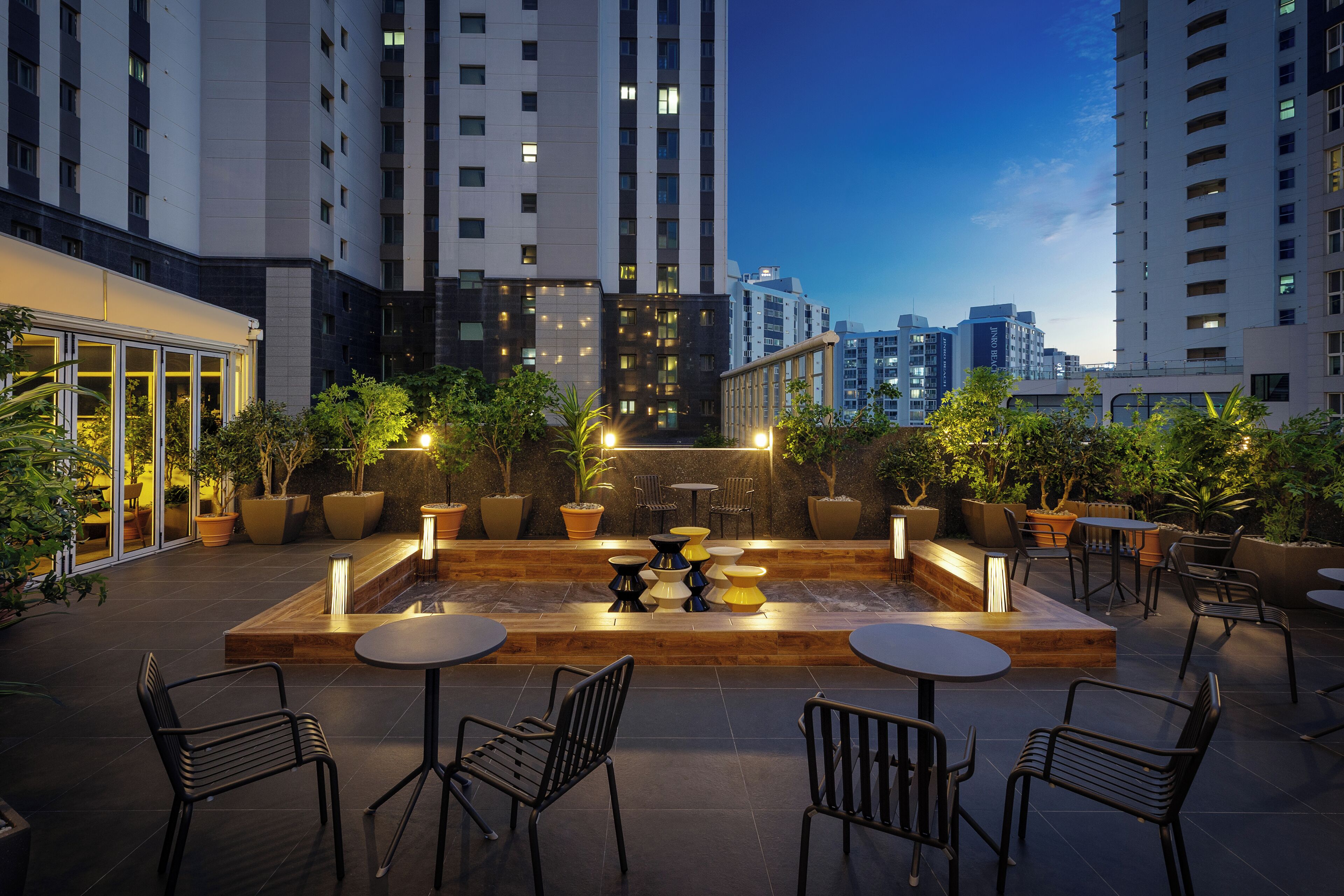 Un espacio al aire libre con mesas y sillas, rodeado de plantas en macetas, con vistas a un paisaje urbano al atardecer.