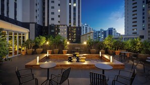An outdoor seating area with tables and chairs, surrounded by potted plants, and overlooking a cityscape at dusk.