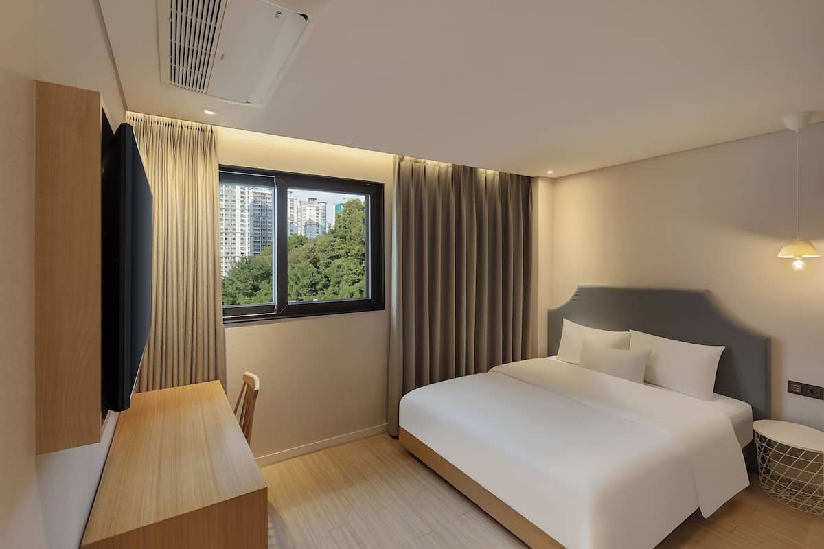 A hotel room with a bed, a desk, a chair, a window with curtains, and a view of buildings and trees.