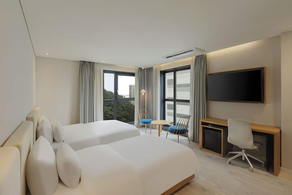 A modern hotel room with two beds, a desk, a chair, a TV, and large windows with curtains.