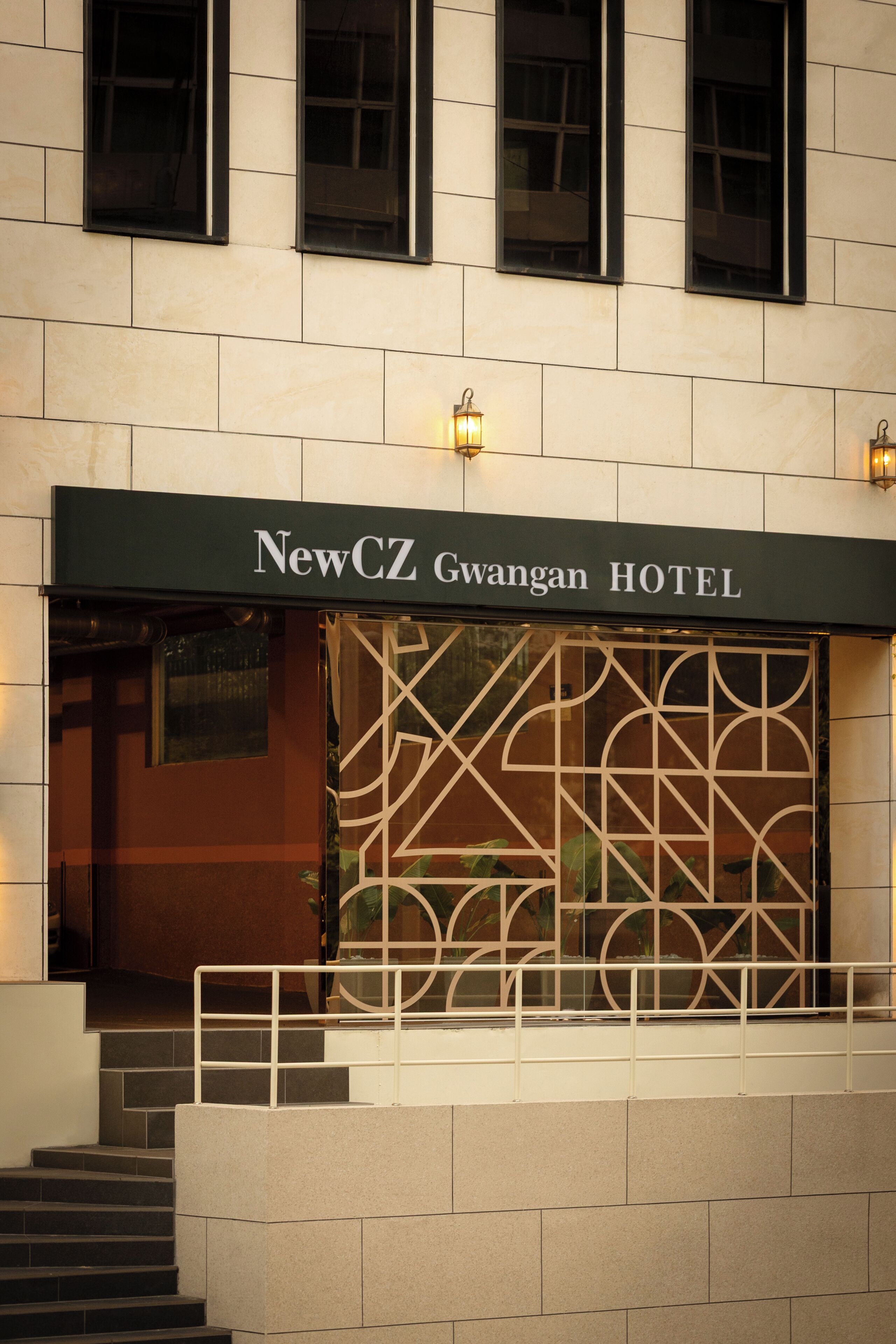 Exterior del Hotel NewCZ Gwangan, con fachada moderna, una entrada destacada y un panel de vidrio decorativo.