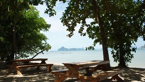 Une plage avec des tables de pique-nique en bois sous les arbres, une étendue de sable et une mer calme où l’on aperçoit des îles au loin.