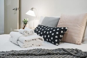 A neatly made bed with folded white towels, a black pillow with white floral patterns, a beige pillow, and a gray knitted blanket. - Corporate Stayz at Collins (Melbourne)