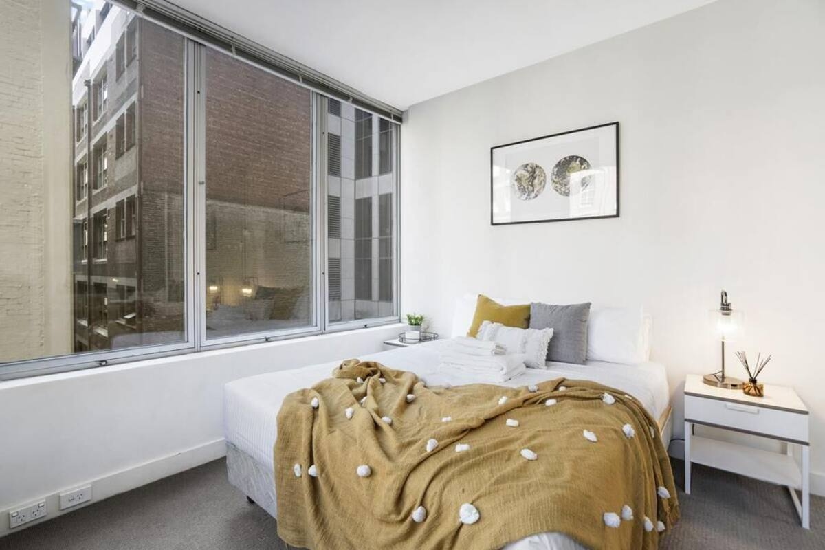 A modern bedroom with a large window, a bed with a white and brown blanket, a bedside table with a lamp, and a framed picture on the wall.