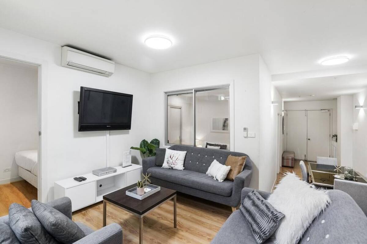 A modern living room with a grey sofa, a coffee table, a flat-screen TV mounted on the wall, and a sliding glass door leading to a dining area.
