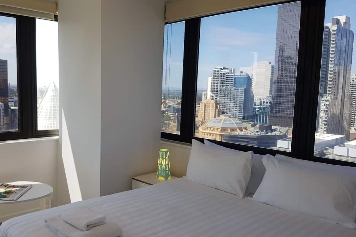 A hotel room with a bed, a small table with a magazine, a lamp, and a view of a cityscape through large windows.