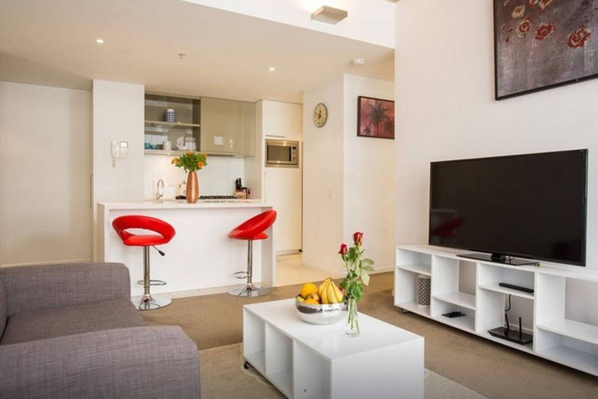 A modern living room with a grey sofa, a white coffee table with fruit and flowers, a TV on a white stand, and a kitchen area with a microwave and red bar stools.