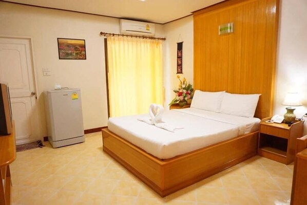 A hotel room with a wooden bed, a small refrigerator, a bedside table with a lamp, and a window with curtains. - Phimaibury Apartment (Phimai)