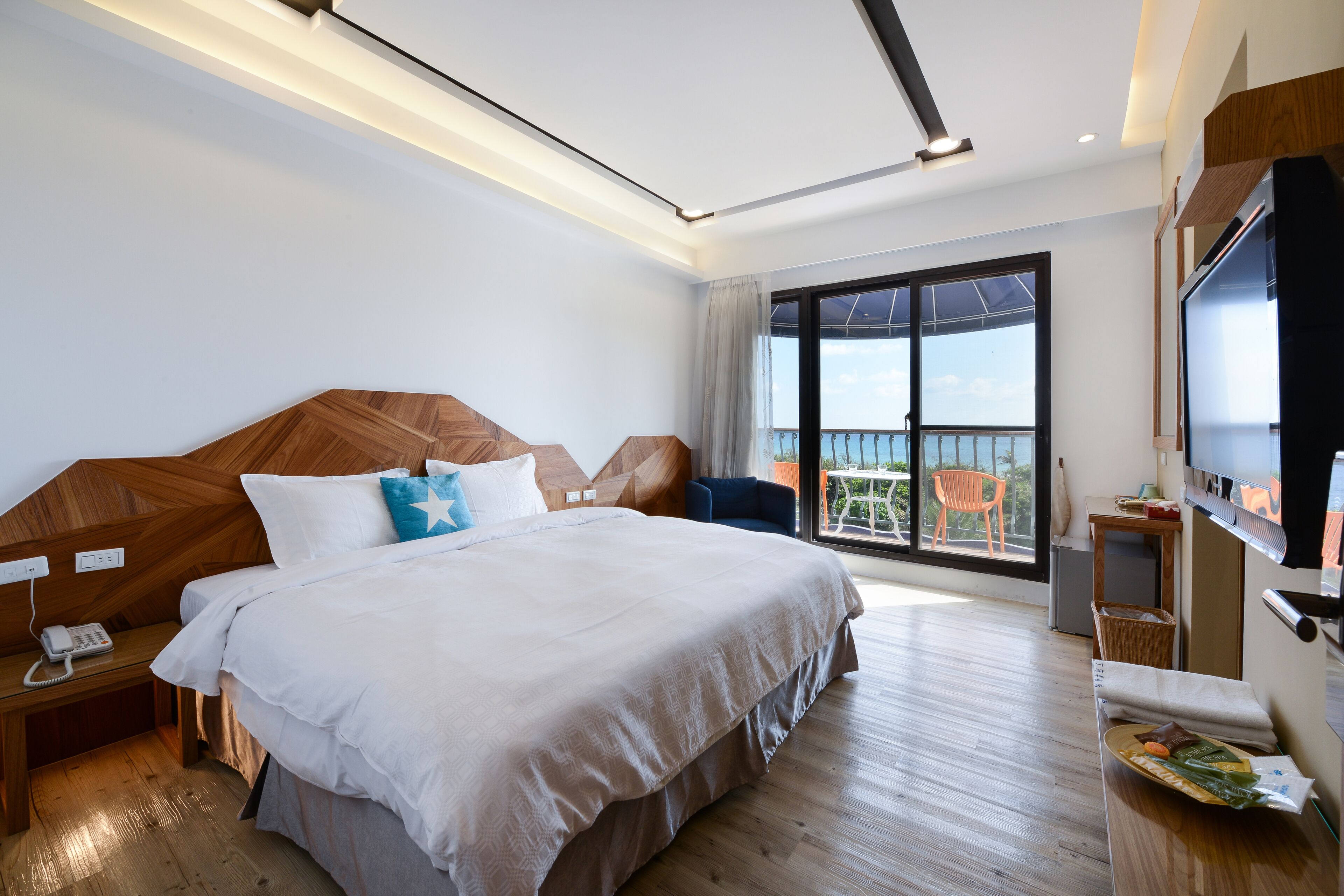 A modern hotel room with a large bed, a TV, a balcony with a chair, and a wooden floor.