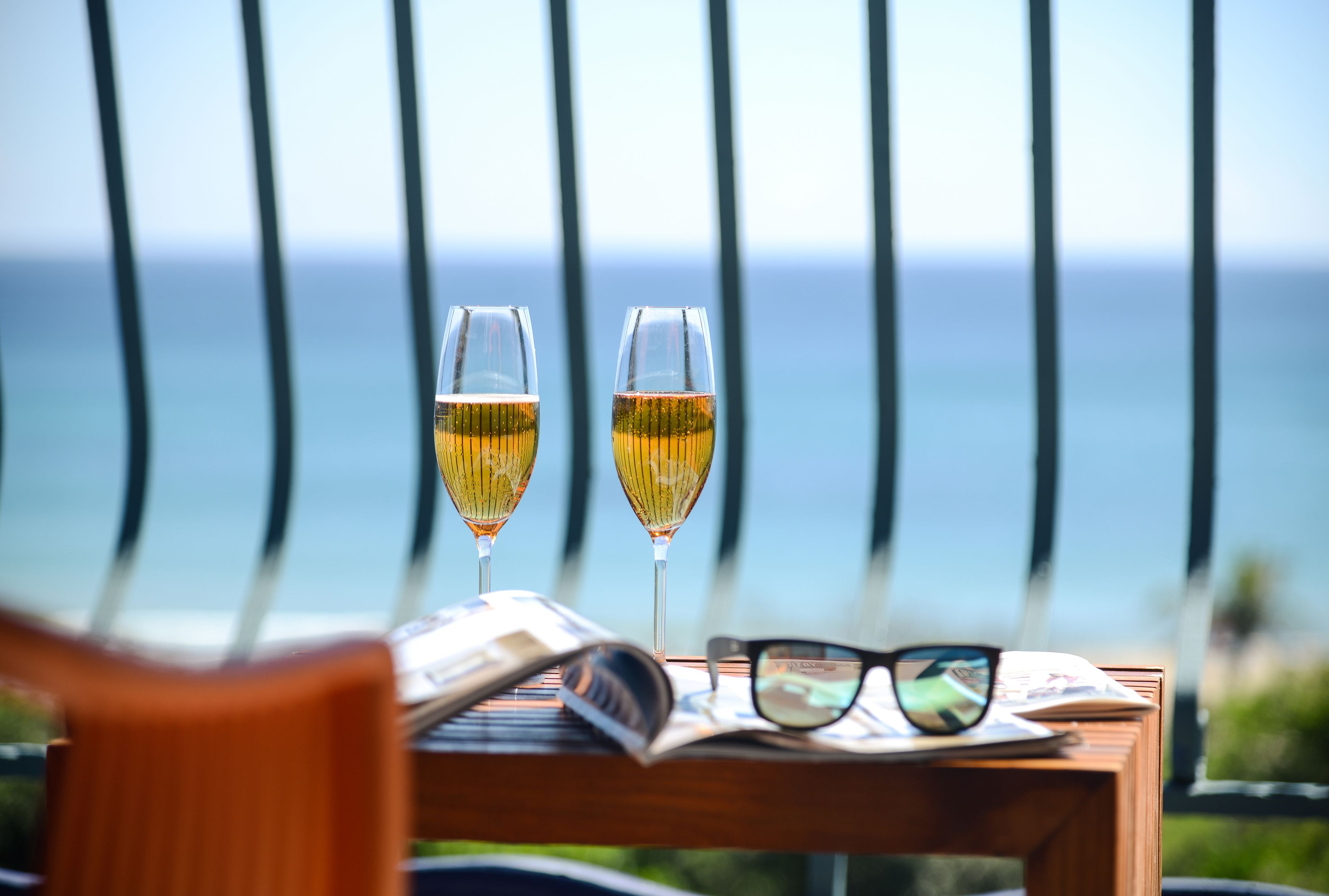 Two glasses of white wine on a wooden table with a magazine and glasses, overlooking a scenic ocean view.