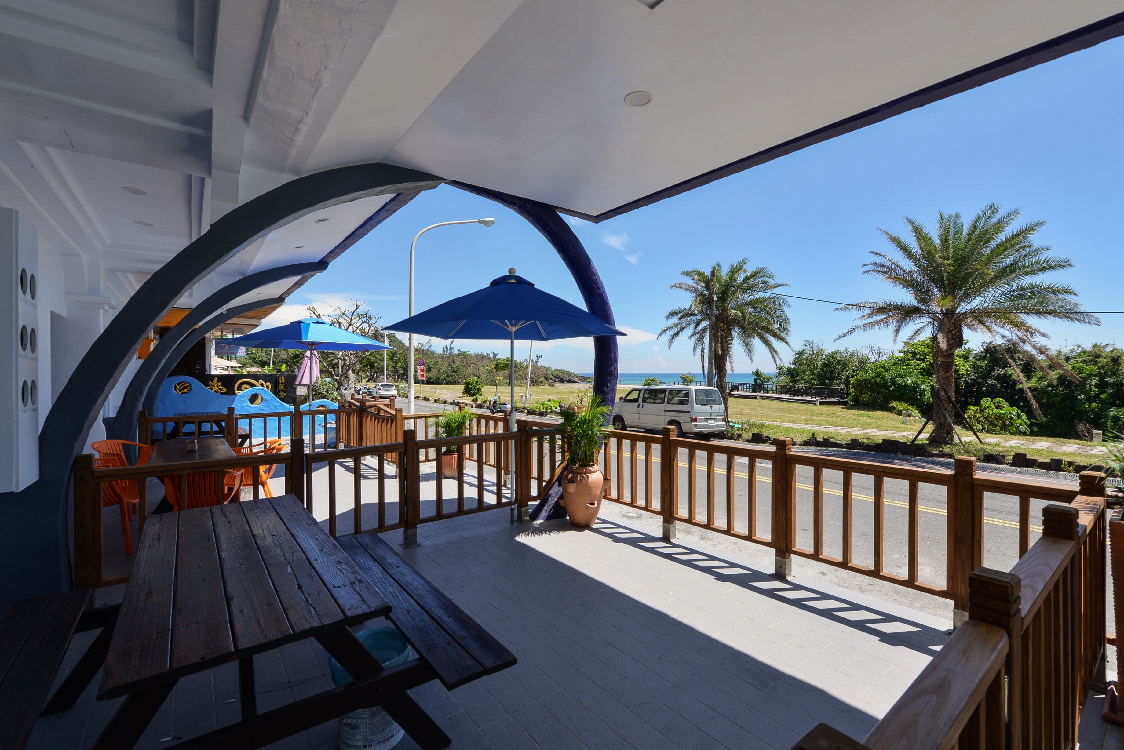 A wooden deck with tables and chairs, a blue umbrella, and a view of the street and palm trees.