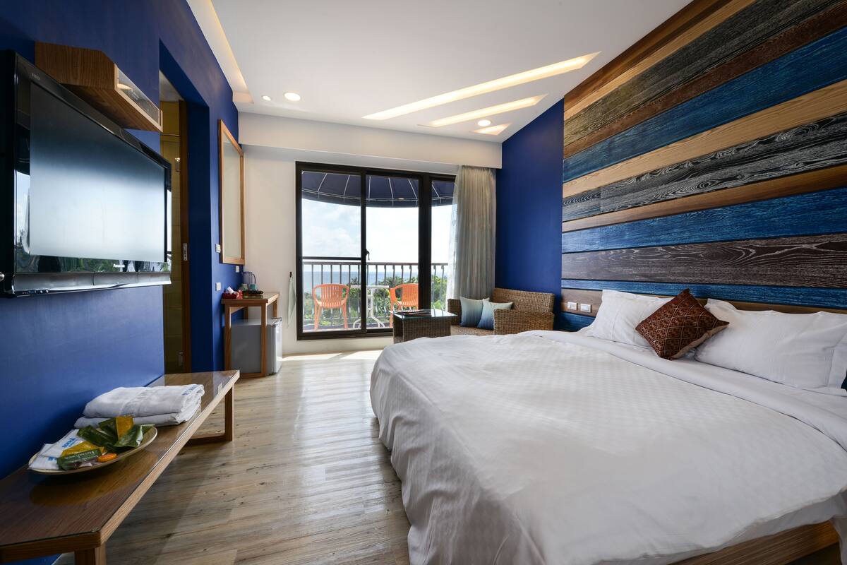 A modern hotel room with a large bed, a flat-screen TV, a balcony with orange chairs, and a wooden headboard.