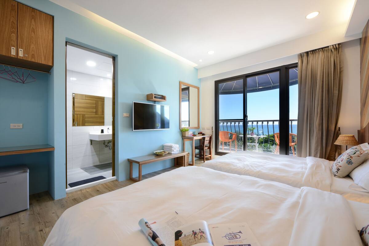 A hotel room with two beds, a TV, a desk, and a balcony with a view.