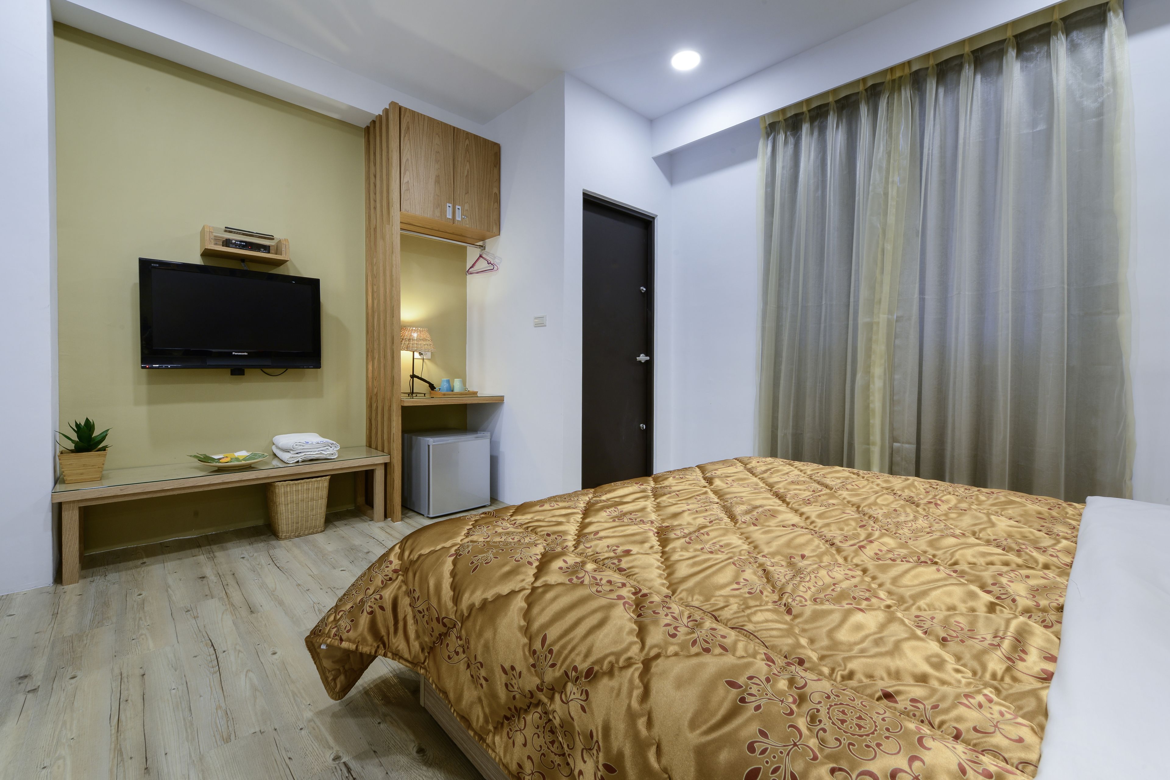 A hotel room with a bed, a television mounted on the wall, a small desk with a lamp, and a door with curtains.