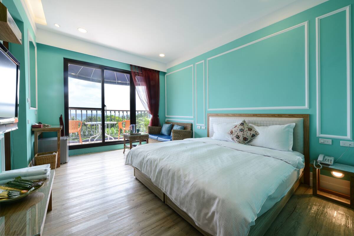 A bedroom with a large bed, a balcony with a view, a small table, and a chair.