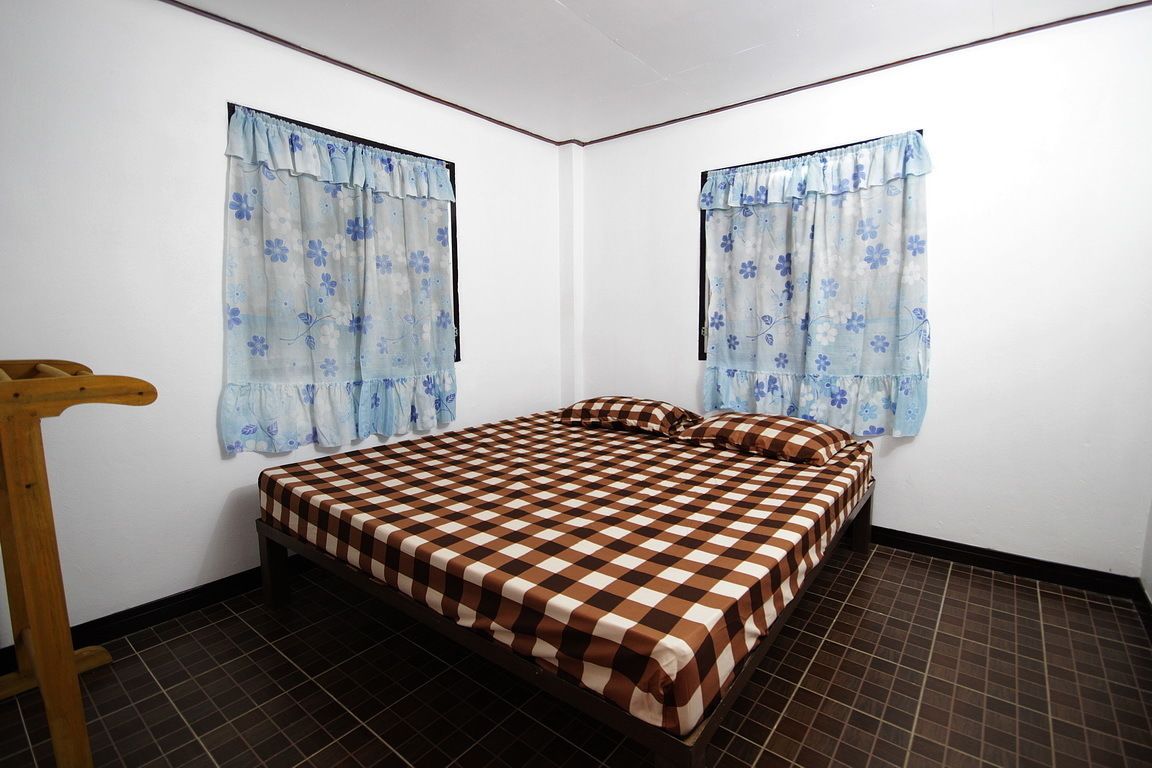Una habitación pequeña con cama individual, colcha de cuadros, dos ventanas con cortinas florales y una silla de madera.
