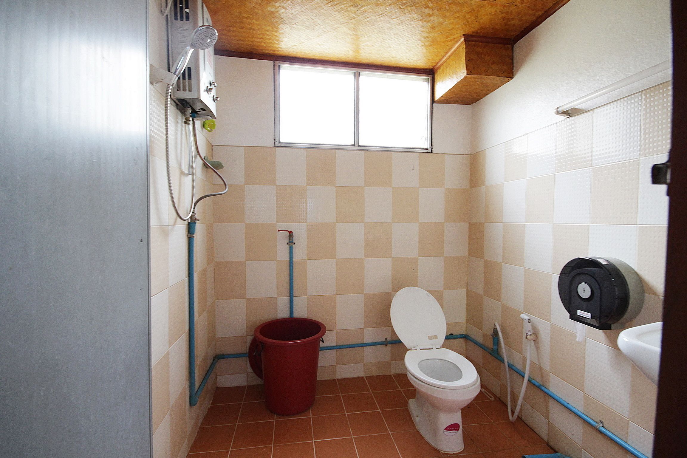 A small bathroom with a toilet, a red bucket, a shower, and a window.