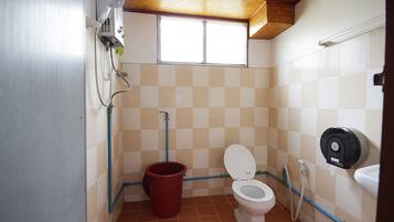 A small bathroom with a toilet, a red bucket, a shower, and a window.