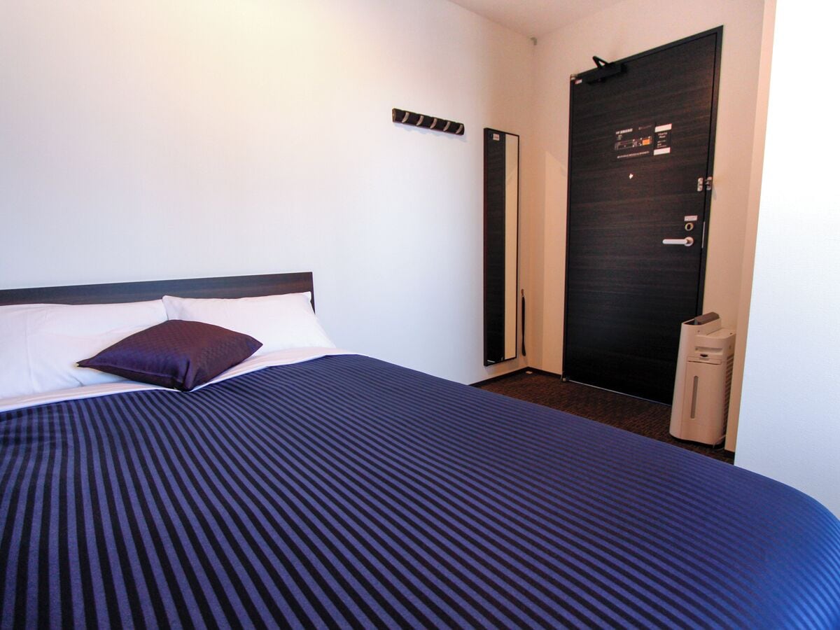 Quarto de hotel com uma cama, uma porta escura, um termostato e uma pequena lixeira.