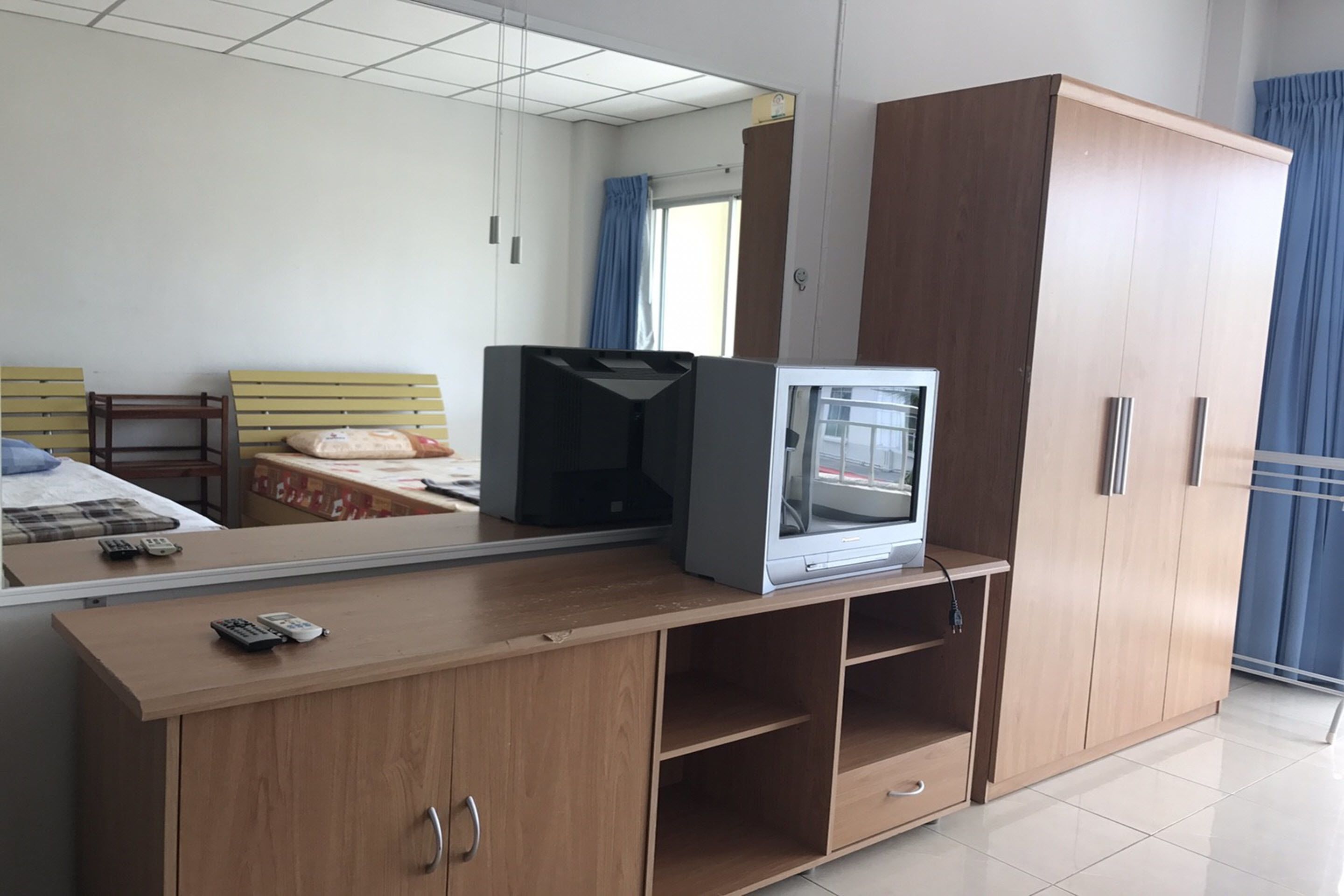 A hotel room with a wooden wardrobe, a bed with a headboard, a television on a wooden stand, and a mirror reflecting the room.