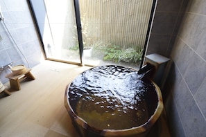 A wooden bathtub with water, a wooden stool, and a window with a view of plants. - KSB Yayoi No Sato Onsen (Izumi)