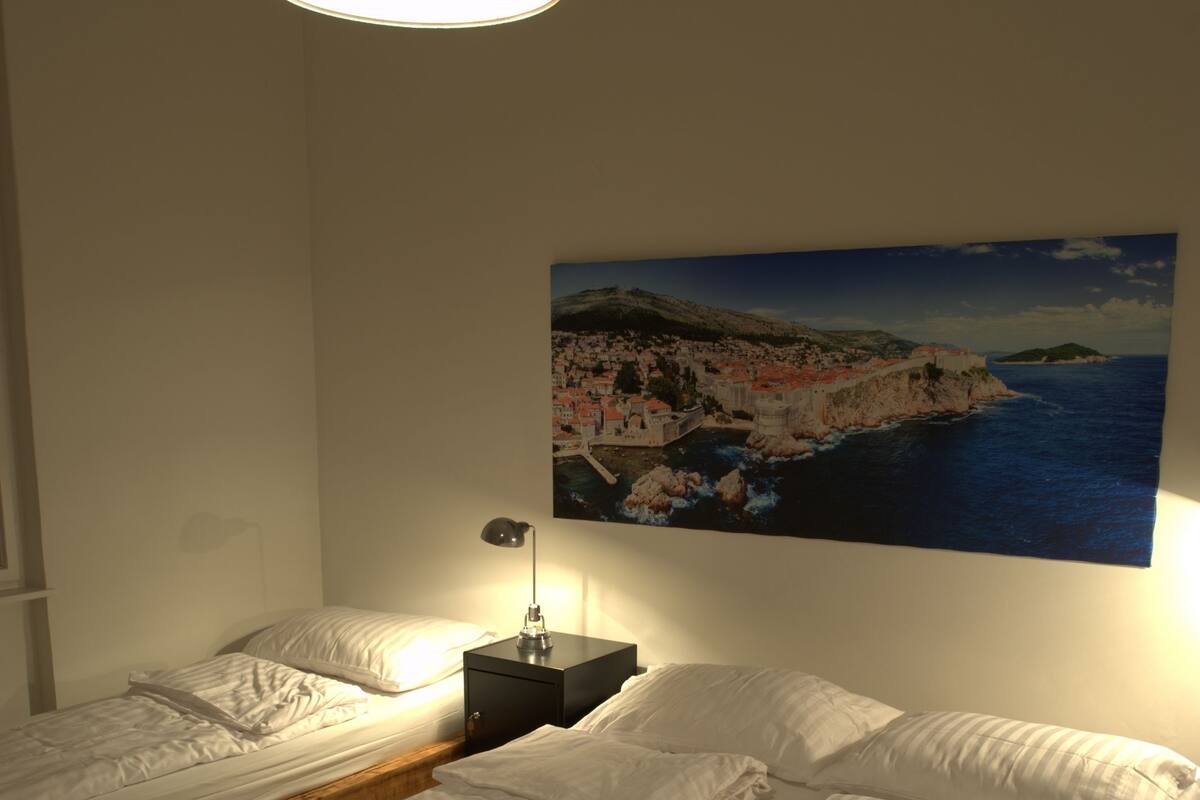 A room with two single beds, a nightstand with a lamp, and a painting of a coastal scene.