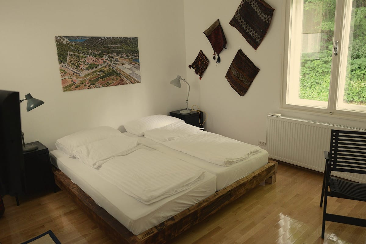 A bedroom with a wooden bed, a nightstand with a lamp, a chair, a window with a view of greenery, and a wall-mounted picture.