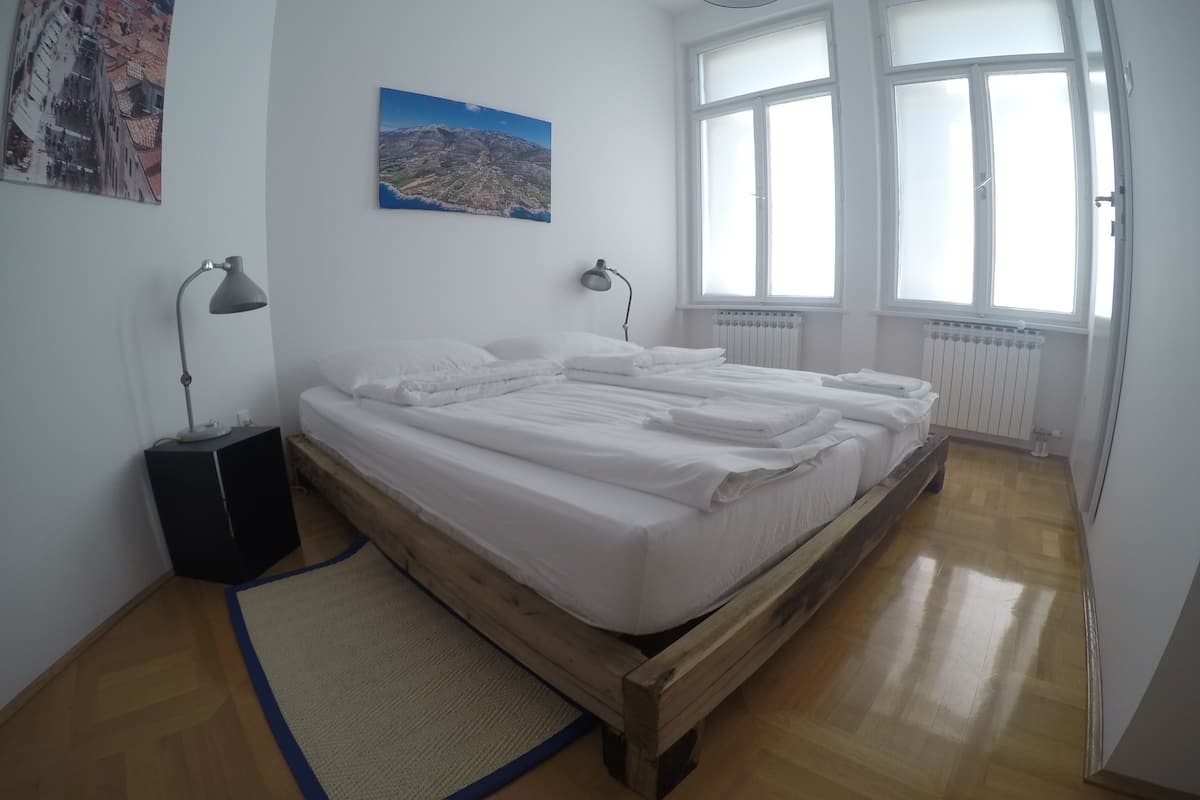 A bedroom with a wooden bed, white bedding, a blue rug, two bedside lamps, a cityscape painting, and a window with blinds.