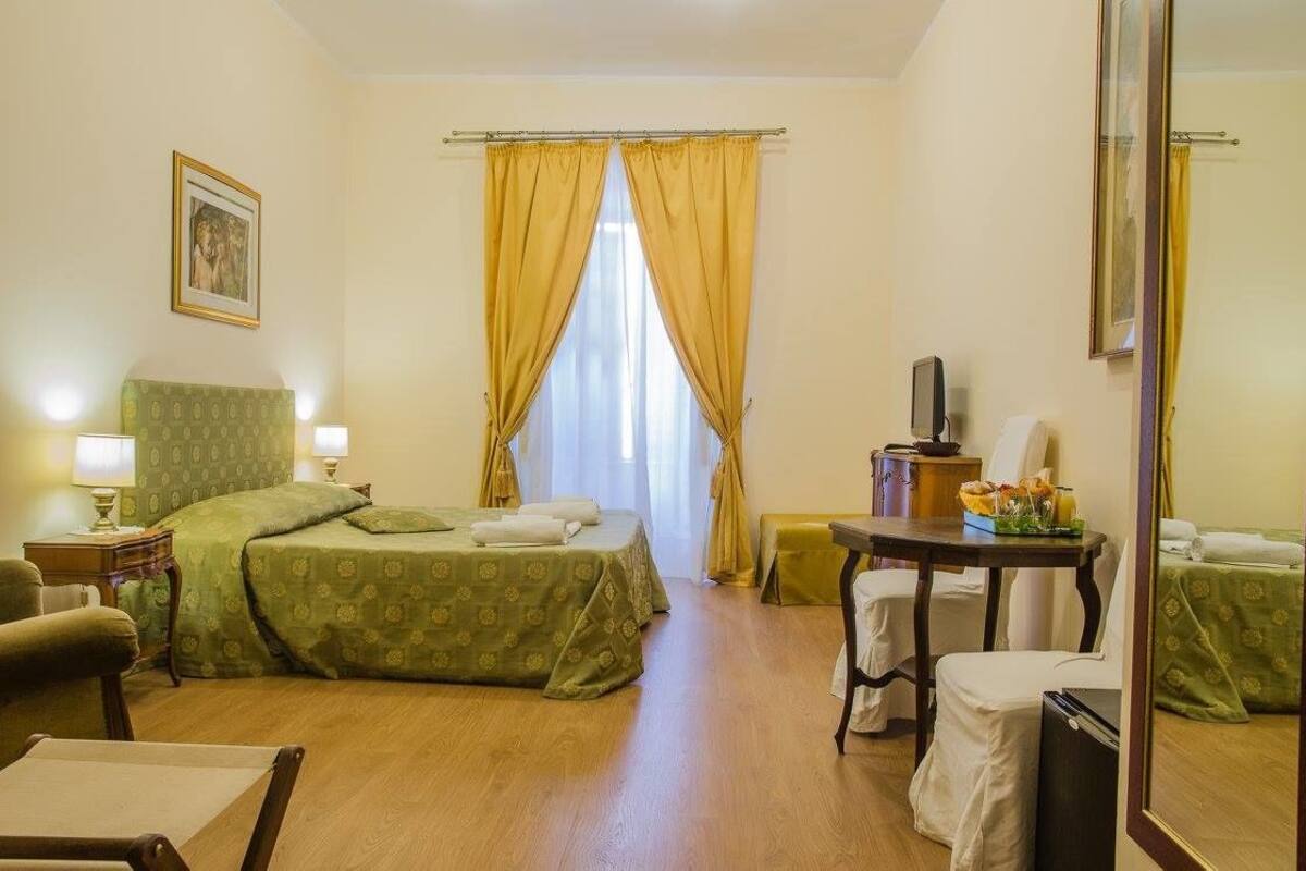 A hotel room with a bed, a desk, a chair, a small table with a vase of flowers, and a mirror.