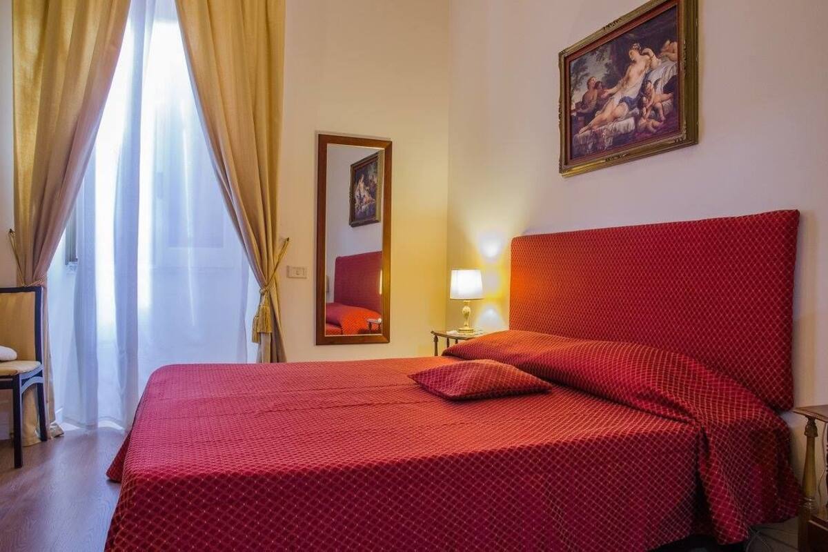 A hotel room with a large bed, a red headboard, a painting on the wall, and a window with curtains.