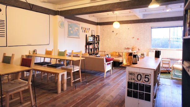 A cozy room with wooden tables and chairs, a whiteboard, and a bookshelf. - Guesthouse SUN - Hostel (Osaka)